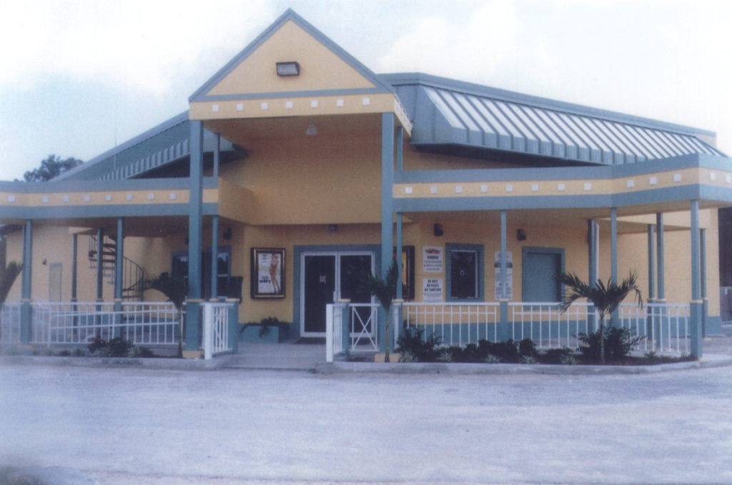 Small commercial building with yellow exterior, teal trim, metal roof, and covered front entrance with white railing.